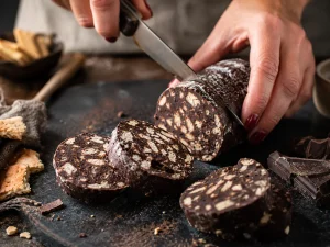 Salam de biscuiți cu cacao, tăiat în felii pe un tocător rustic, alături de biscuiți și bucăți de ciocolată.